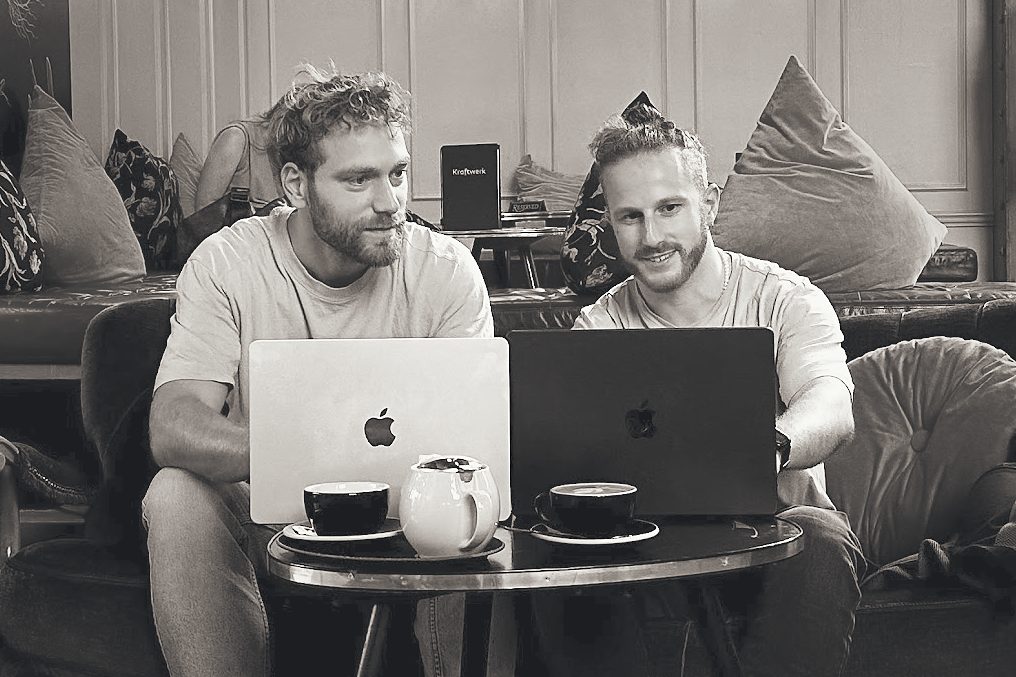 Two people at a small café table with laptops and coffee cups, looking at the same screen